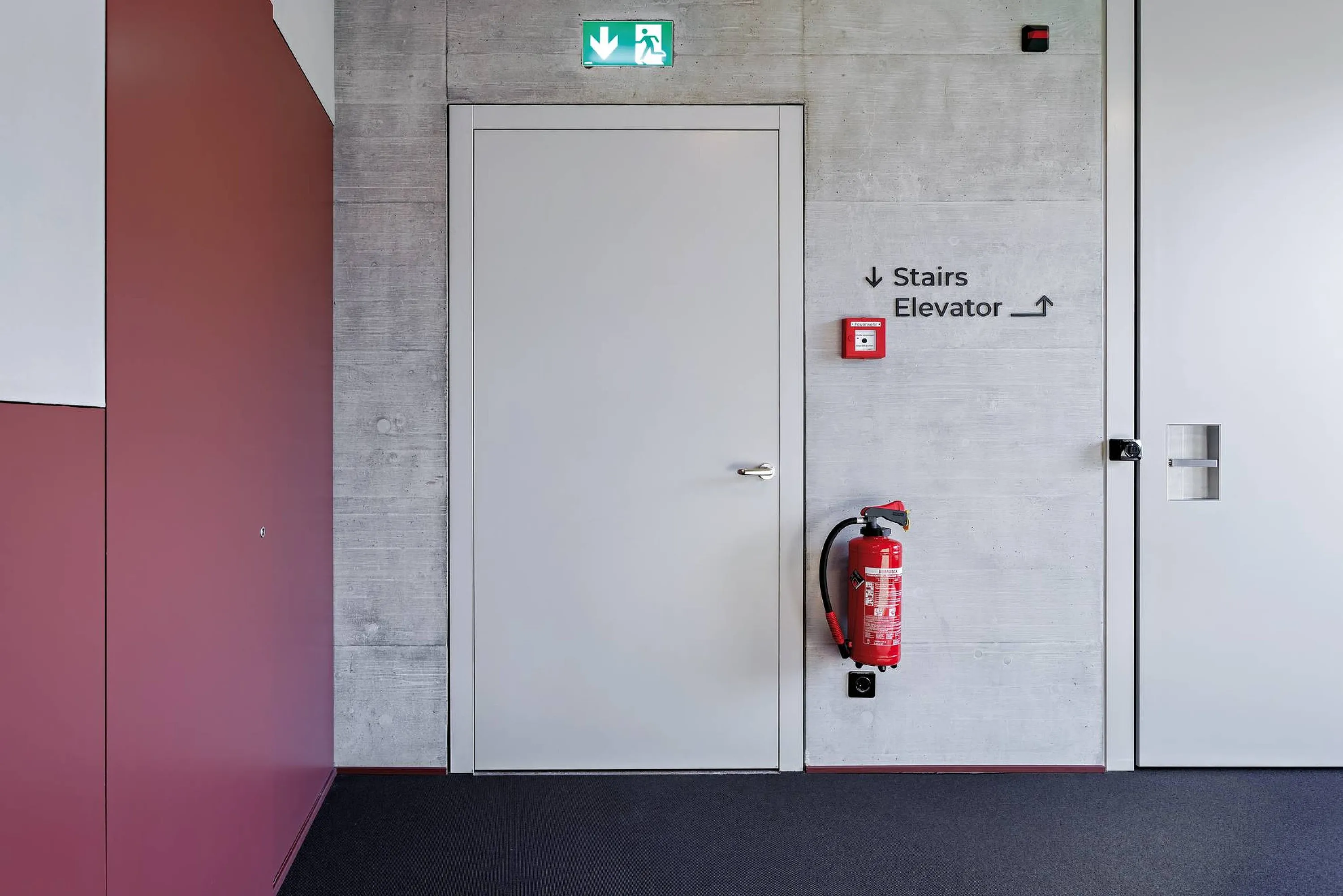 Porte intérieure gris clair avec huisserie sobre et poignée métallique dans le couloir de l'hôtel. Au-dessus, symbole vert de la sortie de secours, à droite, sur le mur en béton, extincteur rouge et alarme incendie, panneau 'Stairs / Elevator' ; à gauche, revêtement mural rouge, moquette sombre.