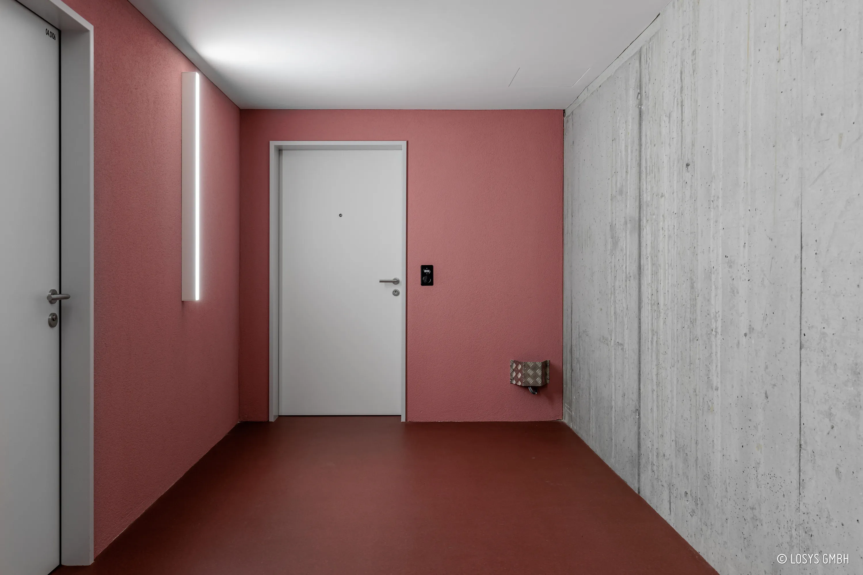 View into a hallway: a white flat entrance door with peephole and handle in a matt red wall in the centre; another white door on the left; raw exposed concrete wall, red-brown floor, vertical LED light and small access module on the right.