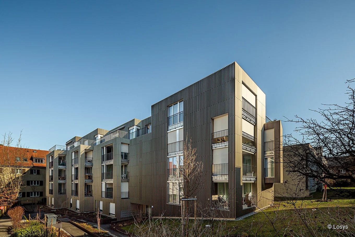 Moderner, viergeschossiger Wohnbau (Alterswohnen WGH St. Katharina, Zürich-Affoltern) bei klarem Himmel: vertikal strukturierte Metallfassade, mehrere Balkone mit Geländern und große Fensterflächen, angelegte Wege und Rasenflächen im Vordergrund, Eingangsbereich an der Gebäudeecke sichtbar; keine Personen zu sehen.