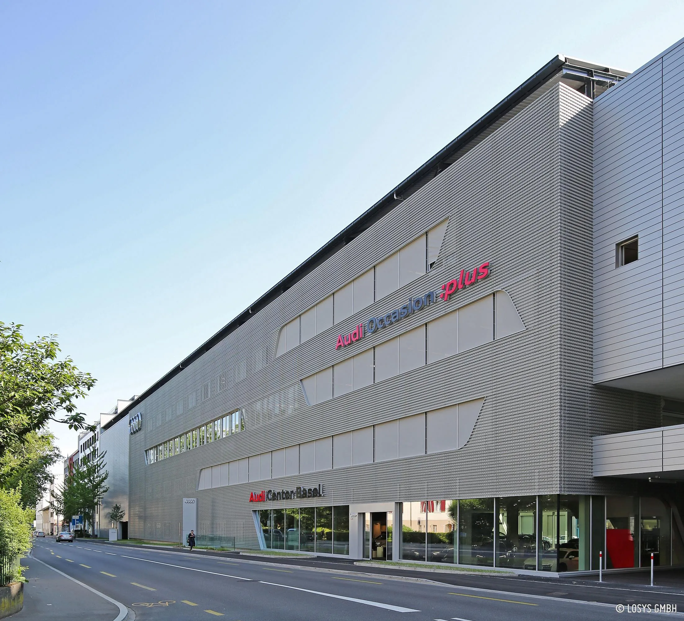 Photo de l'extérieur d'un centre Audi moderne : long bâtiment de plusieurs étages avec une façade métallique gris clair, de grandes vitrines en verre et des inscriptions Audi visibles ; rue, piste cyclable et arbres au premier plan.