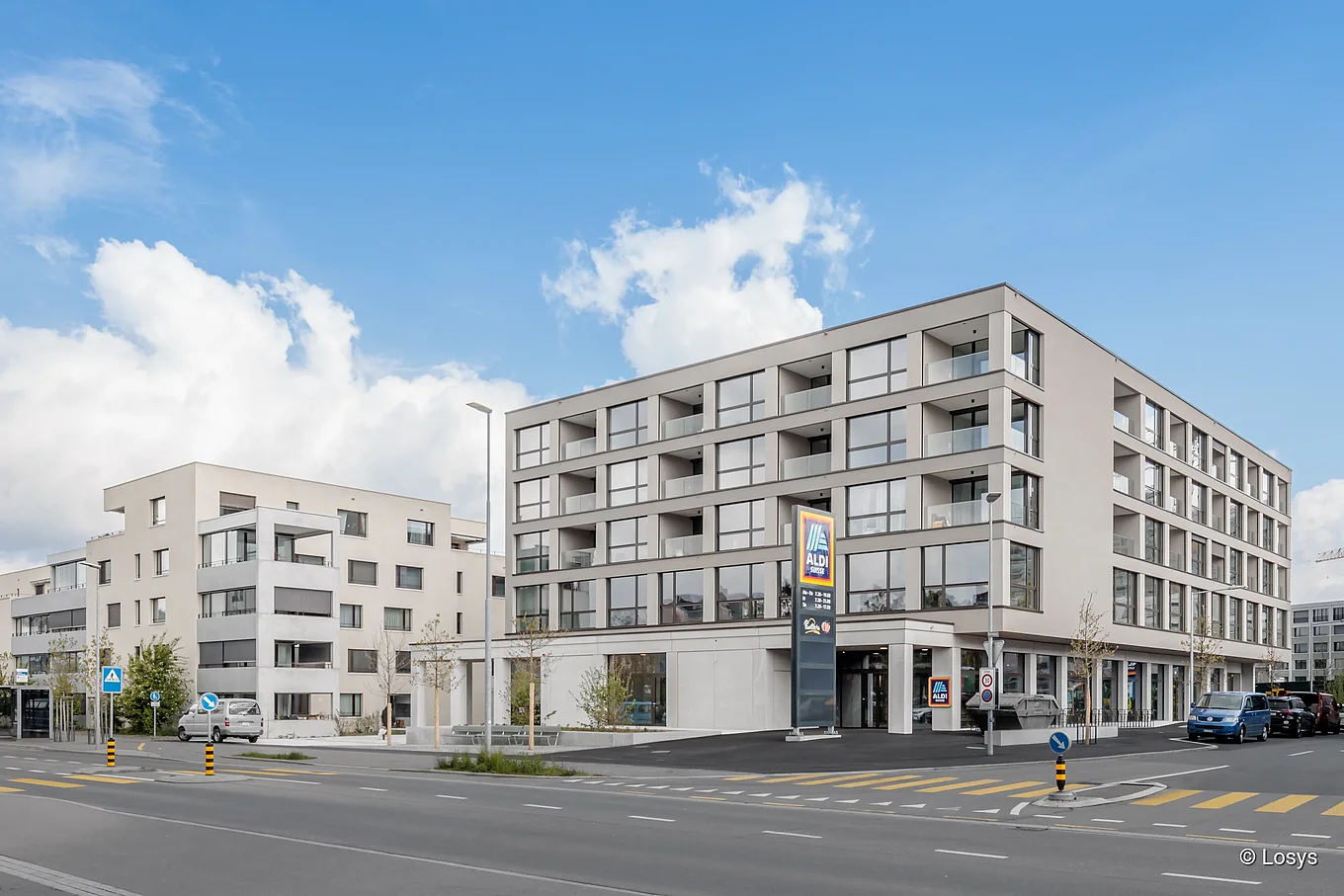 Gebäudefront eines modernen fünfgeschossigen Wohn‑ und Geschäftshauses an einer Straßenecke: Erdgeschoss mit Aldi‑Filiale, darüber großflächige Glasfenster und Balkone, helle Putzfassade, Fußgängerüberweg, parkende Autos und blauer Himmel.