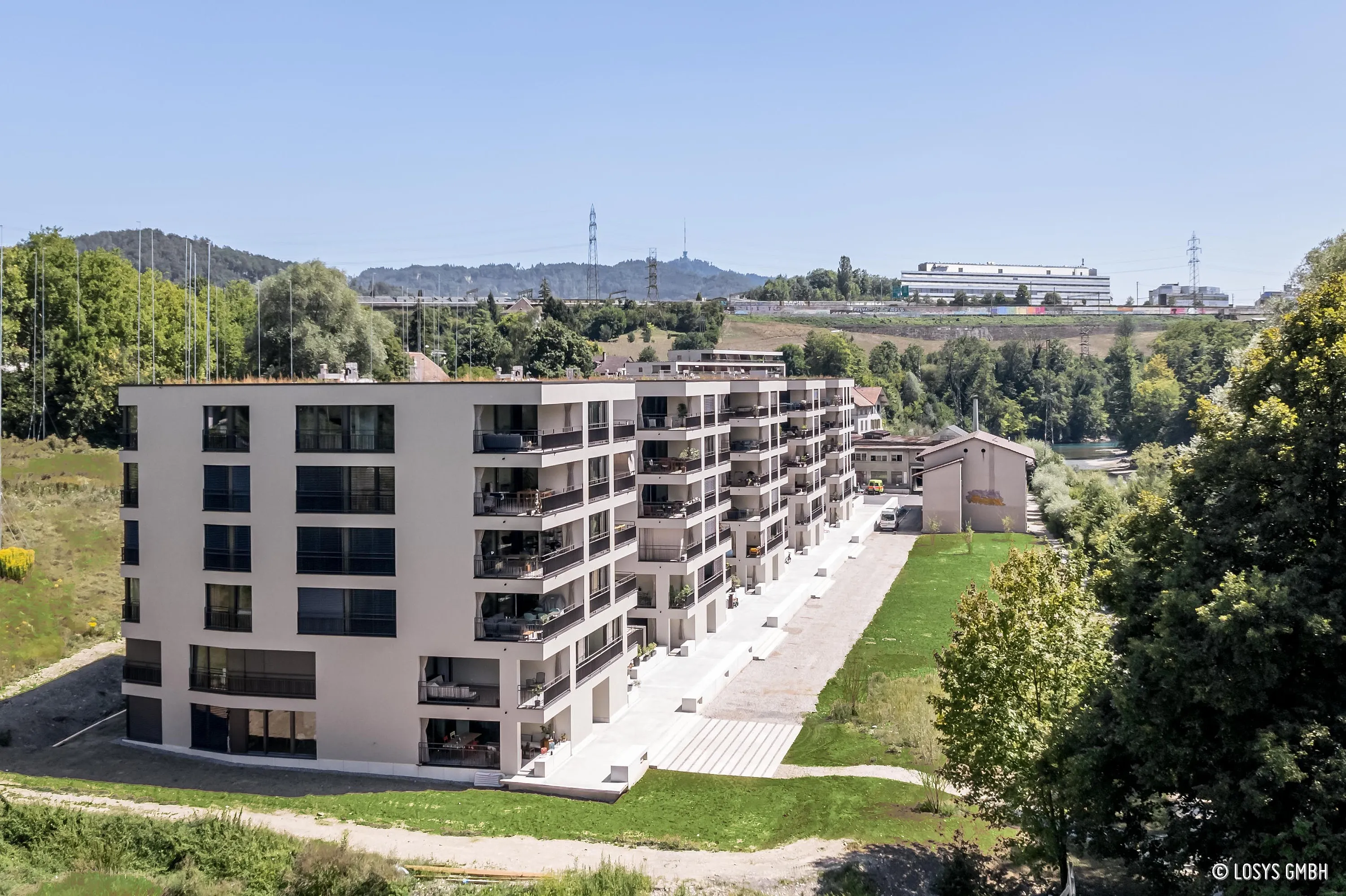 Multi-storey residential building on the banks of the River Aare with continuous balconies, large window fronts and sliding glass doors; surrounding meadows, trees, path and river in the background.