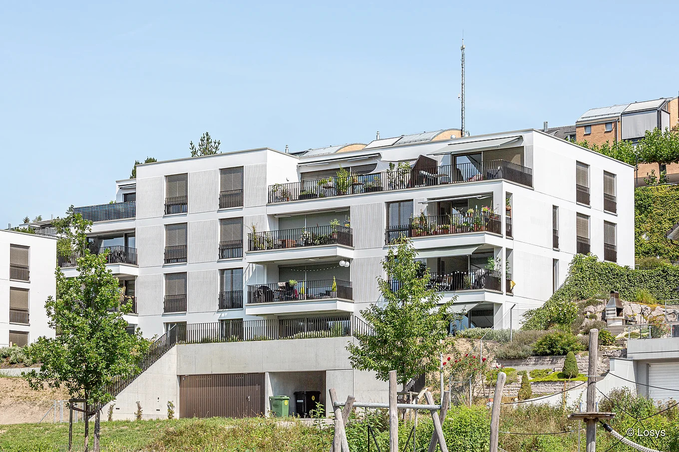 Außenansicht eines modernen, weiß verputzten Mehrfamilienhauses in Hanglage: vier Geschosse mit breiten Balkonen und Terrassen, metallischen Brüstungen sowie großen bodentiefen Fenstern und Glasschiebetüren; Sockel mit Garagentoren, angelegte Grünflächen und junge Bäume im Vordergrund. Keine Personen sichtbar.