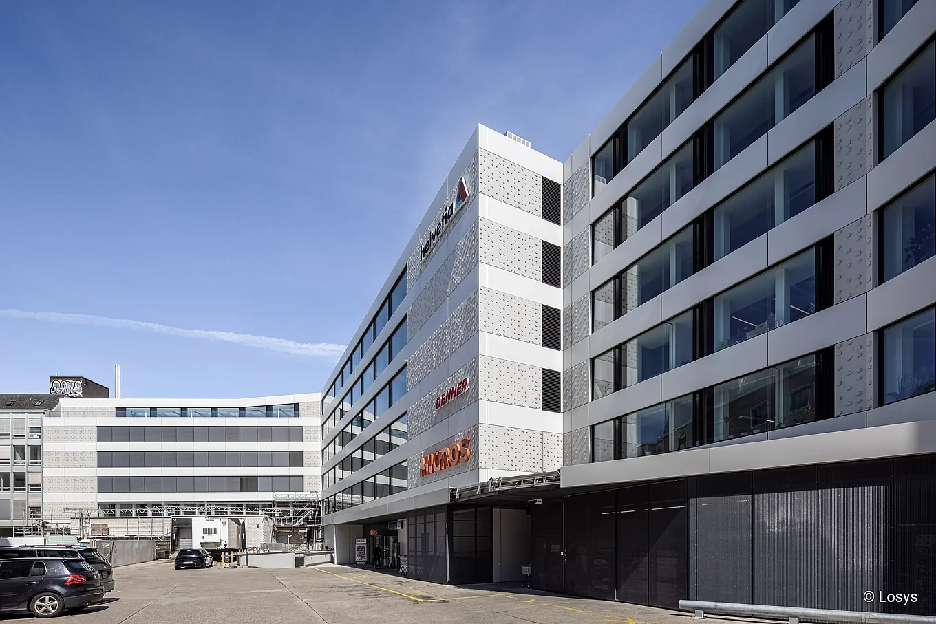 Außenansicht des Geschäftshauses Wengihof in Zürich: Innenhof zwischen zwei Gebäudeflügeln, moderne weiße Fassaden mit horizontalen Bändern und großen Glasfenstern, dunkel abgesetztes Erdgeschoss mit Eingangsüberdachung, sichtbares "Denner"-Logo und Park-/Ladezone im Vordergrund.
