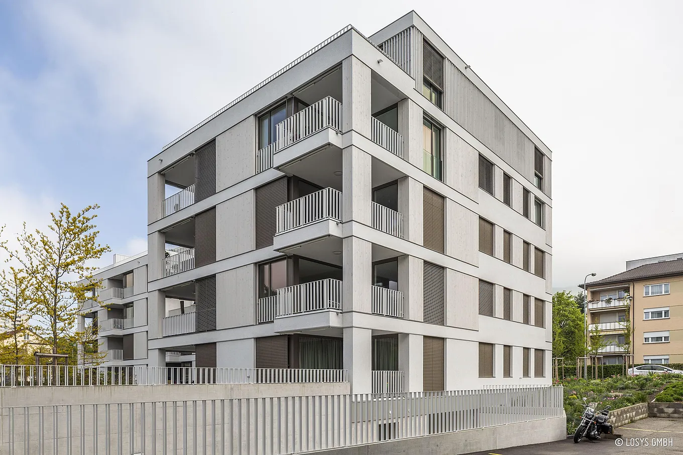 Eckansicht eines modernen viergeschossigen Wohngebäudes: helle Betonfassade mit zurückversetzten Balkonen und vertikalen Geländern, geschlossene Rollläden und großflächige Glas‑Balkontüren; im Vordergrund Zaun, Bepflanzung und ein geparktes Motorrad.