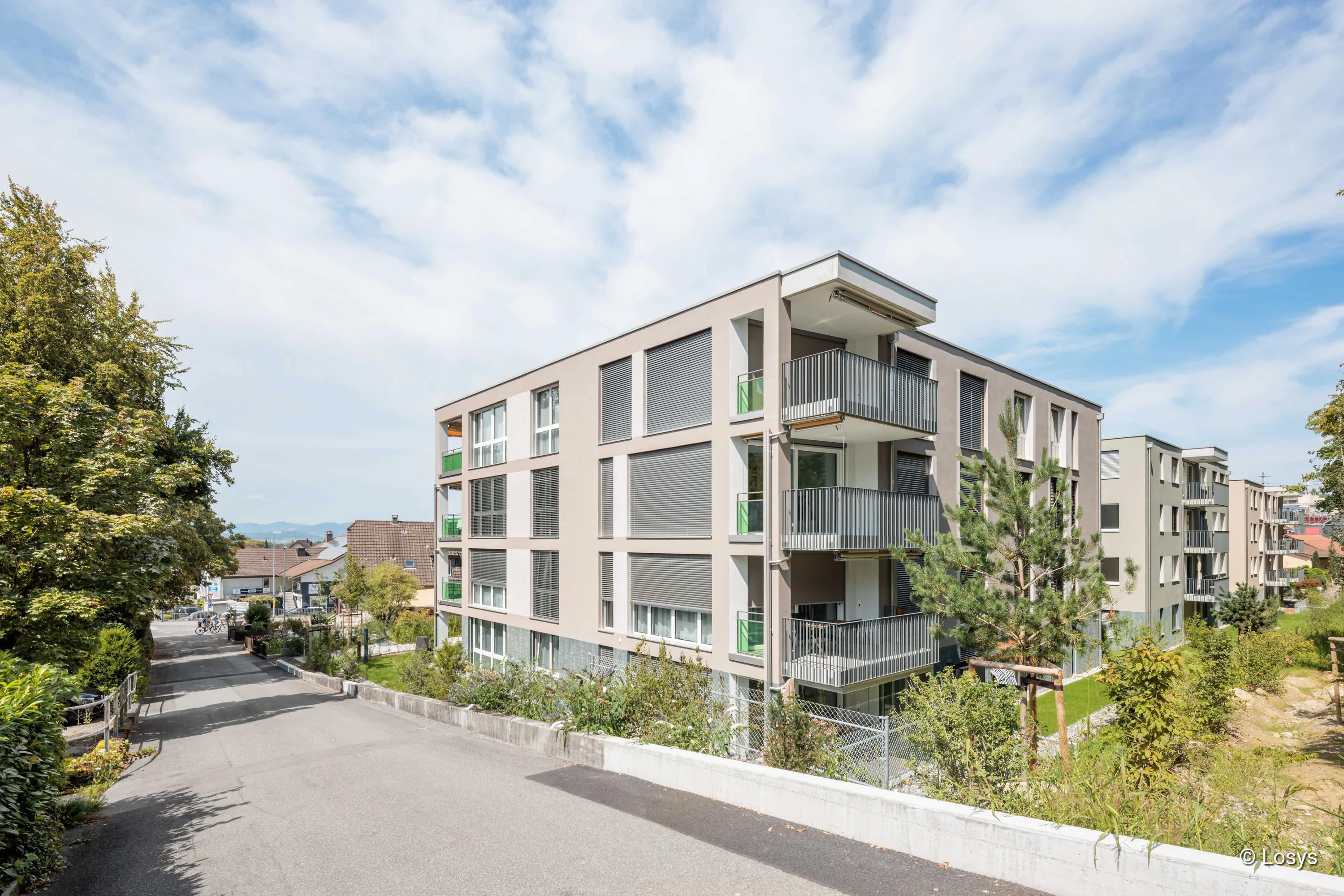 Eckiges, viergeschossiges Mehrfamilienhaus mit heller Putzfassade, grauen Rollläden und Balkonen mit dunklen Metallgeländern; Vorgarten mit Bäumen, angrenzende Wohnstraße und Nebengebäude sichtbar.