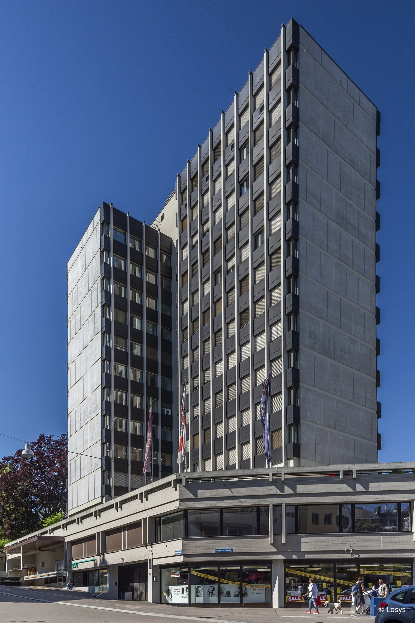 Vorderansicht des Bauhof-Gebäudes in Zürich-Oerlikon: zwei nebeneinanderstehende hohe Beton- und Glas­türme mit regelmässigen dunklen Fensterrahmen über einem flachen, zweigeschossigen Sockel mit Schaufenstern und Fahnen; Personen auf dem Gehweg.