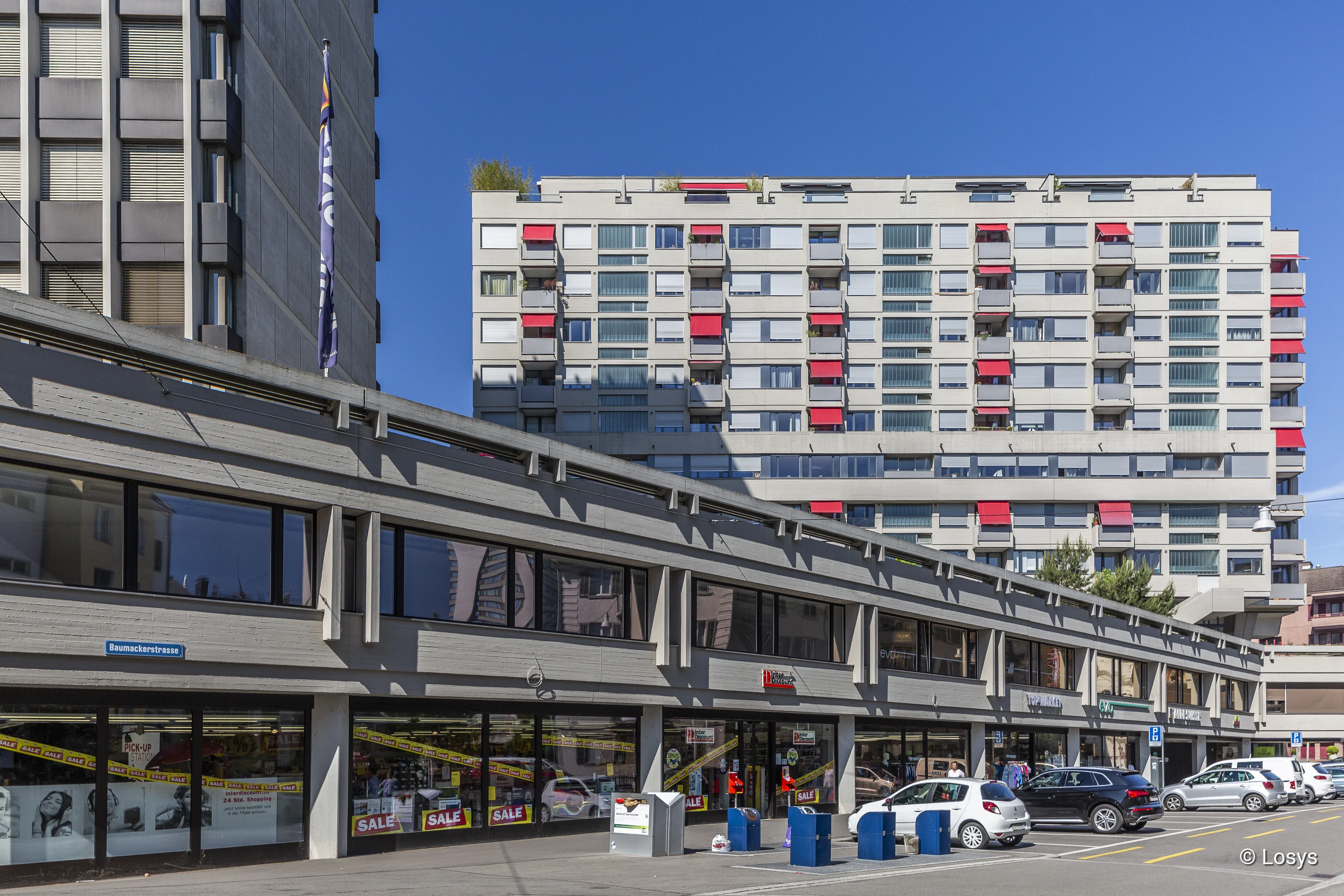 Vordergrund: lange zweigeschossige Beton-Ladenzeile mit großen Schaufenstern und SALE-Schildern, davor parkende Autos; im Hintergrund ein mehrstöckiges Wohnhaus mit weißen Fassaden und roten Balkonmarkisen, blauer Himmel.