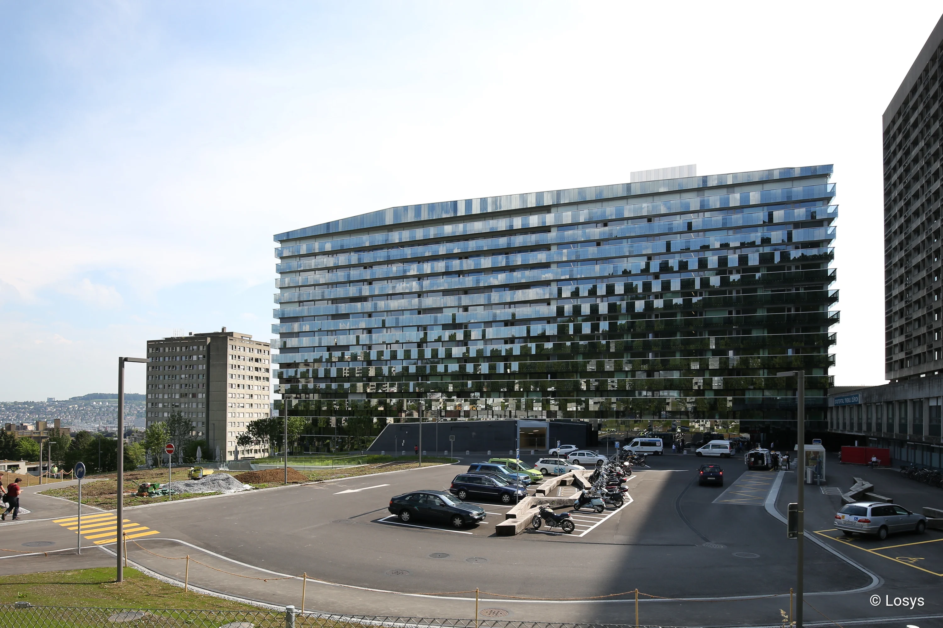 Bettenhaus des Triemli‑Spitals in Zürich: breiter, mehrstöckiger Gebäuderiegel mit spiegelnder Glasfassade in horizontalen Bändern. Im Vordergrund großer Vorplatz mit Parkflächen, markierten Zufahrten, mehreren parkenden Autos und Motorrädern; rechts und links angrenzende Hochhäuser. Aufnahme bei Tageslicht vom Straßenniveau.