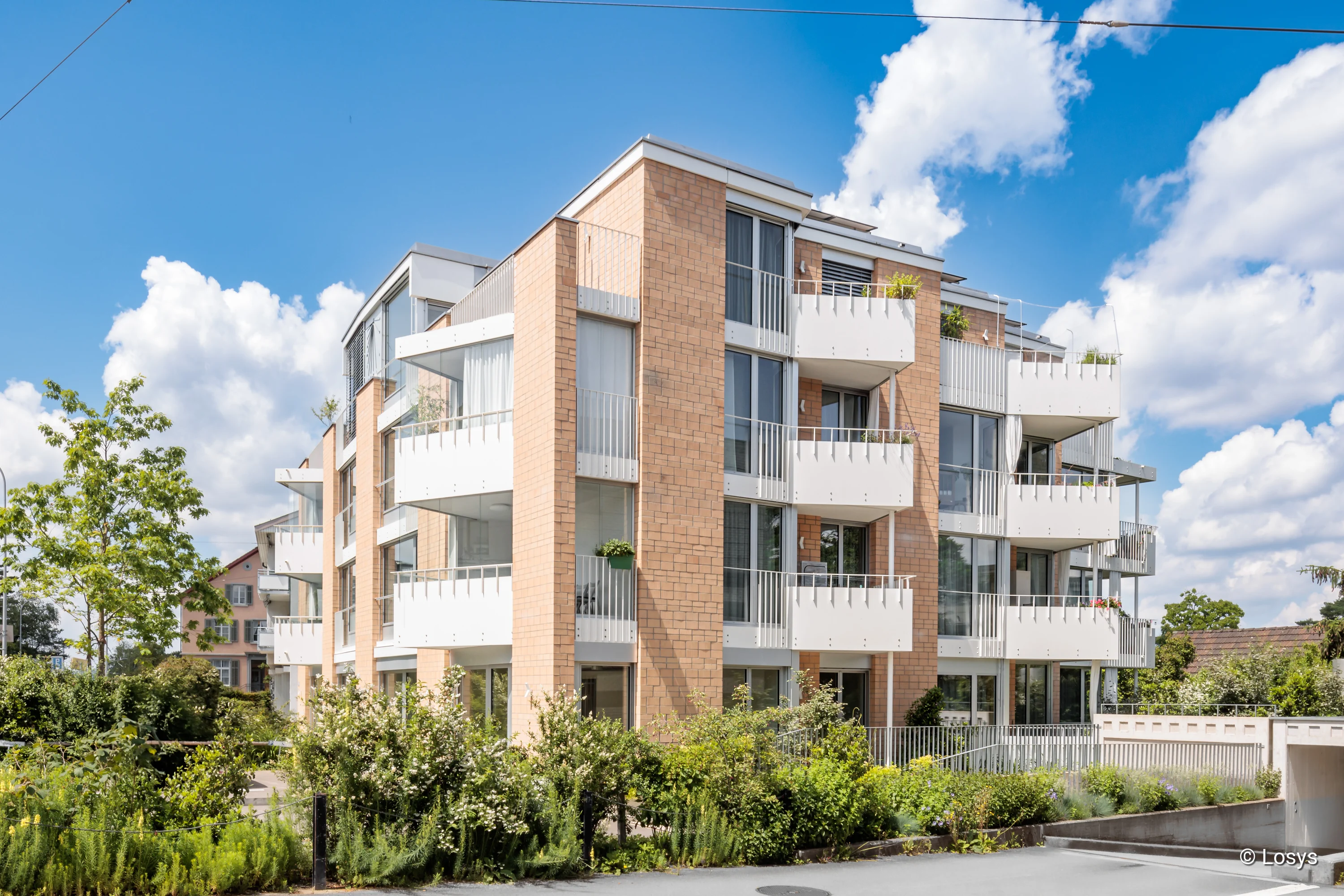 Außenansicht eines modernen Mehrfamilienhauses mit hellen Klinkerfassaden, bodentiefen Fenstern/Glasschiebetüren, weißen Balkonen, begrünter Umgebung und Zufahrt zur Tiefgarage.