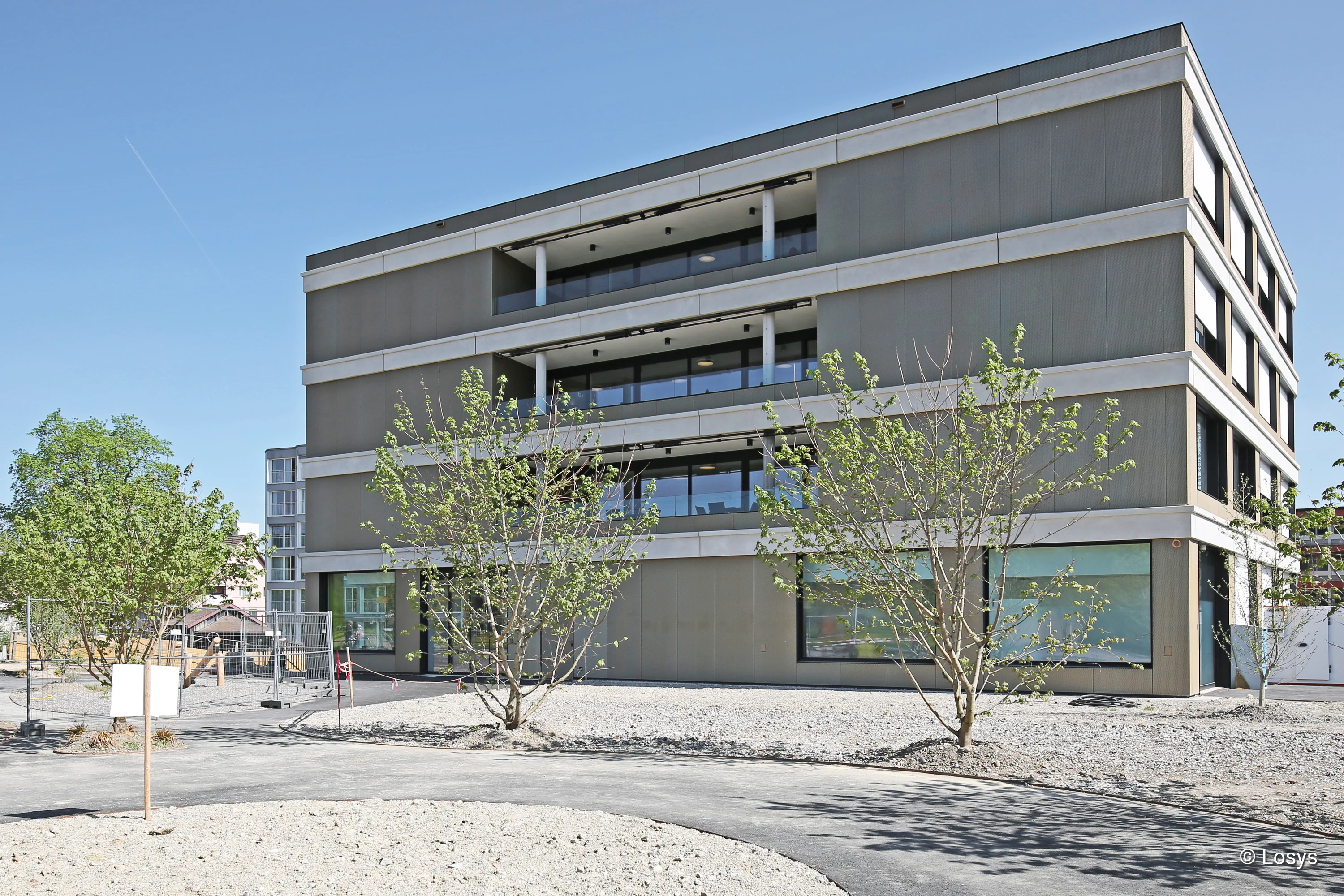 Viergeschossiger, moderner Erweiterungsbau eines Alters- und Pflegezentrums mit horizontal gegliederter Fassade, großen Glasfenstern und zurückgesetzten Balkonen; im Vordergrund Kiesfläche, junge Bäume, Fußweg und Bauzaun.