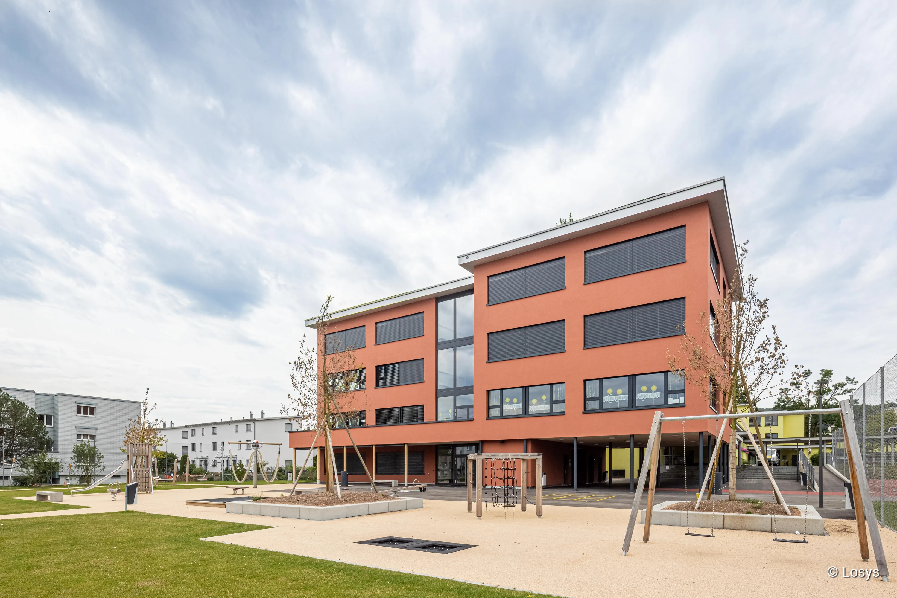 Mehrstöckige Erweiterung der Schulanlage Höegler in Dübendorf: modernes Gebäude mit orange-roter Putzfassade und zentraler vertikaler Glaszone, großen rechteckigen Fenstern mit dunklen Sonnenschutzläden. Im Vordergrund ein offener Pausenhof mit Rasenflächen, gepflasterten Wegen, jungen Bäumen, Schaukeln, Klettergerüst und einem überdachten Durchgang unter einem zurückspringenden Gebäudeteil; bewölkter Himmel.