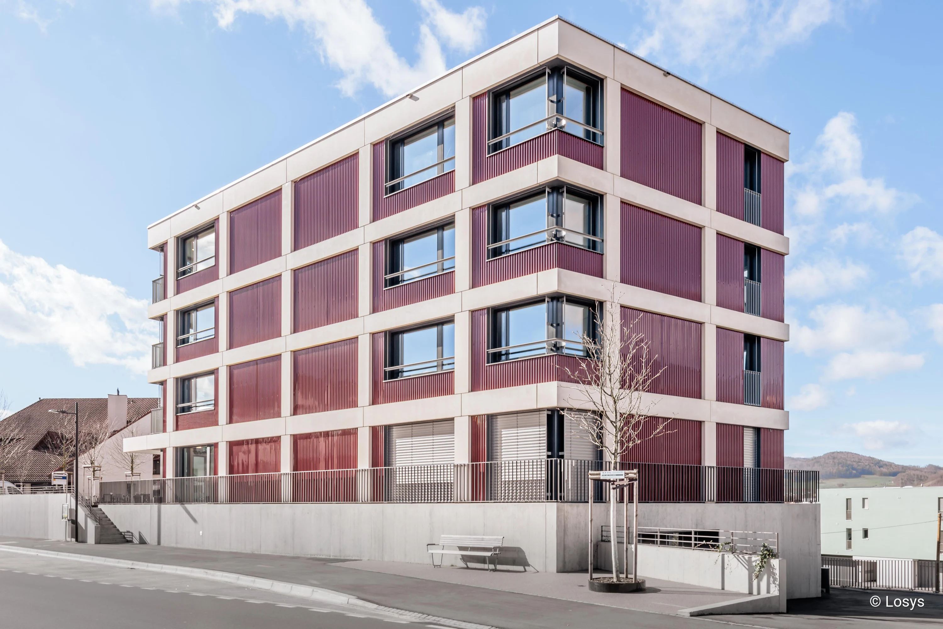 Viergeschossiges Wohnhaus an Straßenecke mit roten Metallpaneelen, Betonbändern, Fenstern, Sockel mit Rampe und Bank.