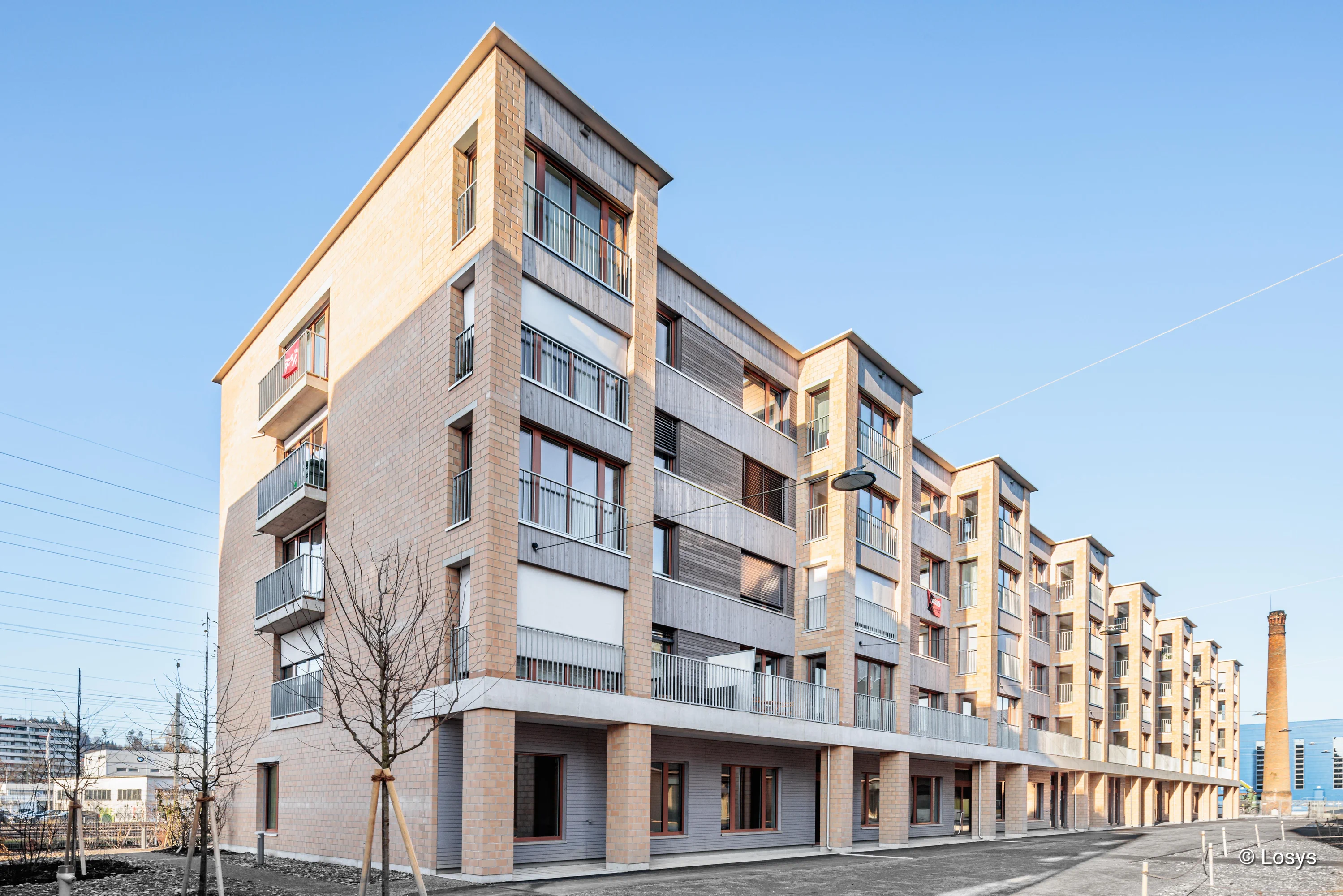 Außenaufnahme: Wohnhaus auf dem Hobelwerk-Areal in Winterthur, fünf Geschosse mit ziegel- und Holzfassade, Reihengalerien/Balkone, bodentiefe Fenster und überdachte Erdgeschosszone.