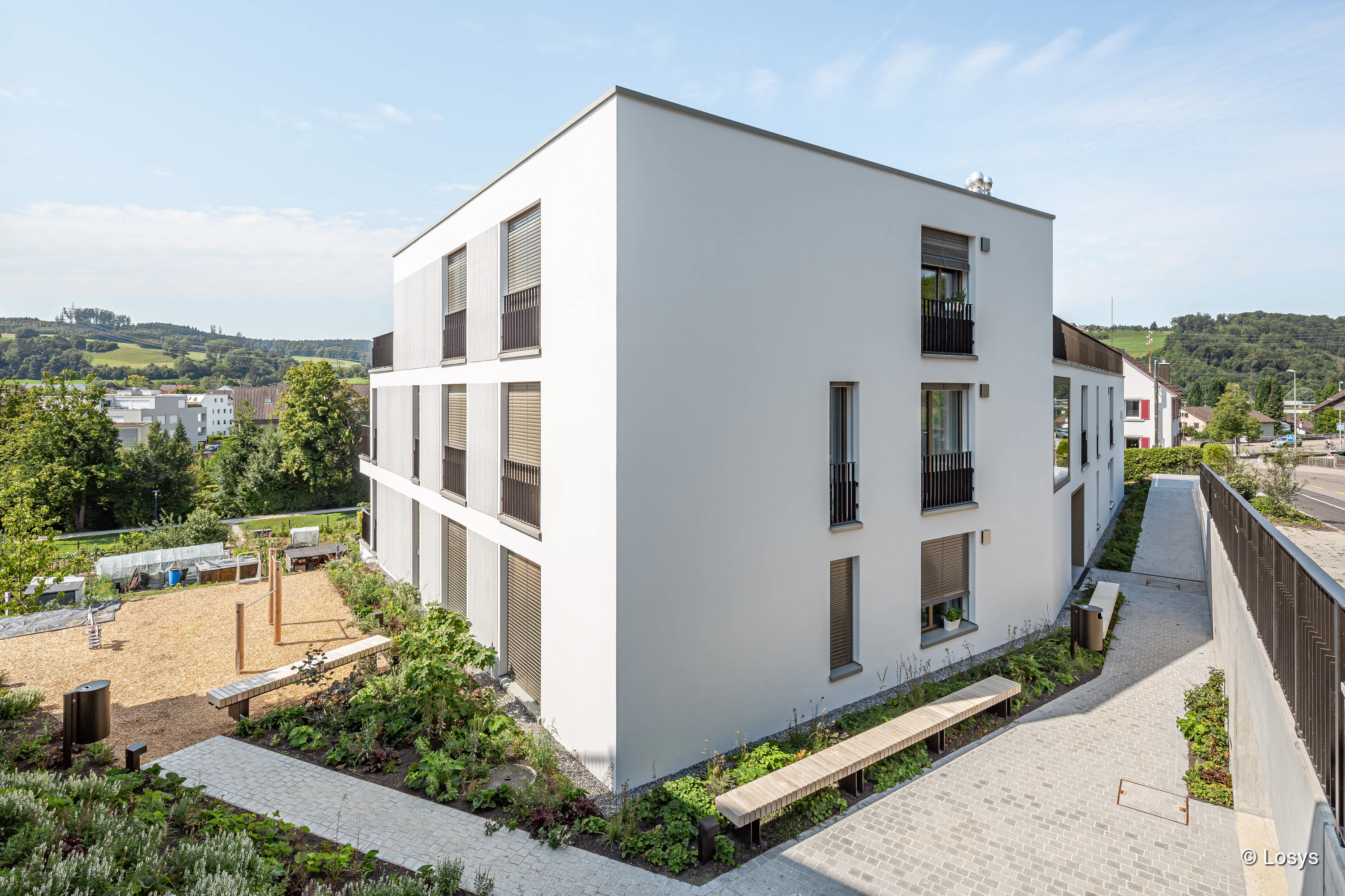 Außenansicht eines modernen, weißen Mehrfamilienhauses (drei Geschosse) mit schmalen Fenstern, außenliegenden Jalousien und kleinen Balkongittern. Gepflasterter Weg mit Sitzbank, angelegte Beete und ein Spielbereich mit Fallschutz; im Hintergrund grüne Hügel und weitere Wohnhäuser. Keine Personen sichtbar.