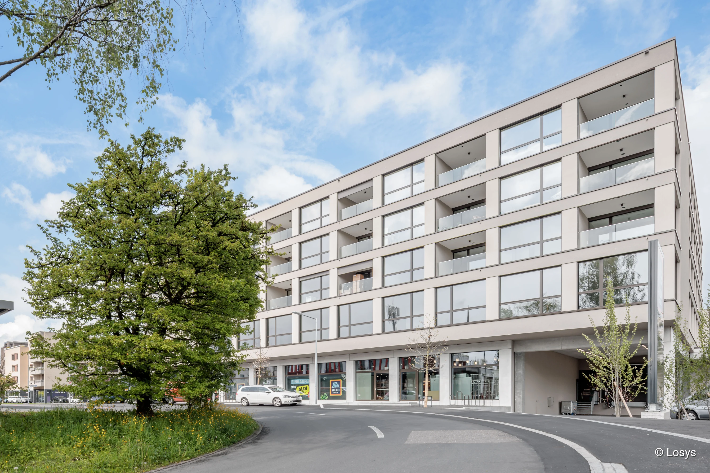 Moderner fünfgeschossiger Neubau mit hellbeiger Fassade und großflächigen Glasfronten; sichtbare Ladenzeile im Erdgeschoss, Balkone auf den Obergeschossen, geschwungene Zufahrt und ein großer Laubbaum im Vordergrund.