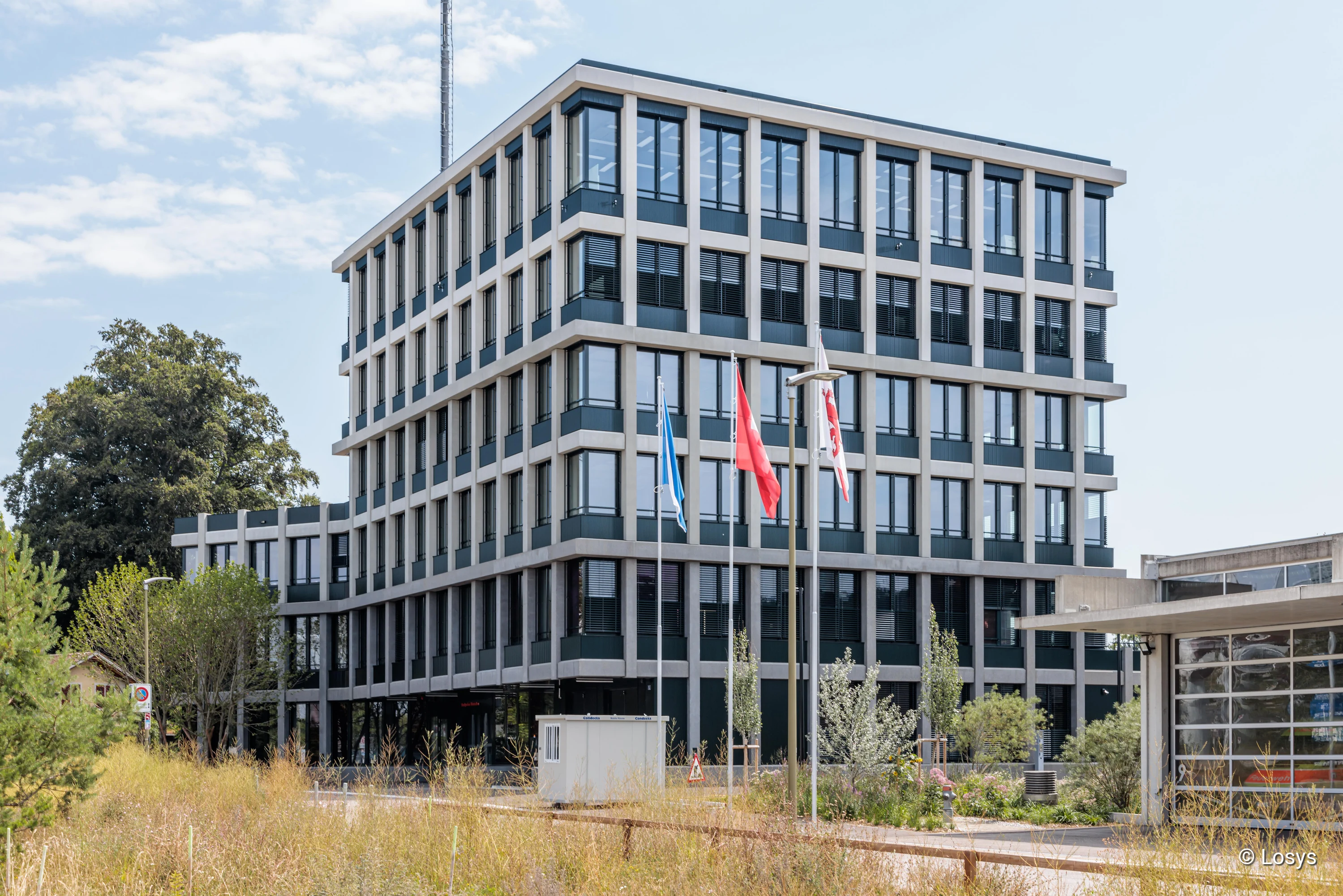 Außenansicht des mehrstöckigen Polizeigebäudes in Winterthur: kubische, regelmäßig gegliederte Fassade mit hellen Betonbändern und großen dunklen Fenstern, drei Fahnenmasten vor dem Eingang, begrüntes Vorfeld und klarer Himmel.