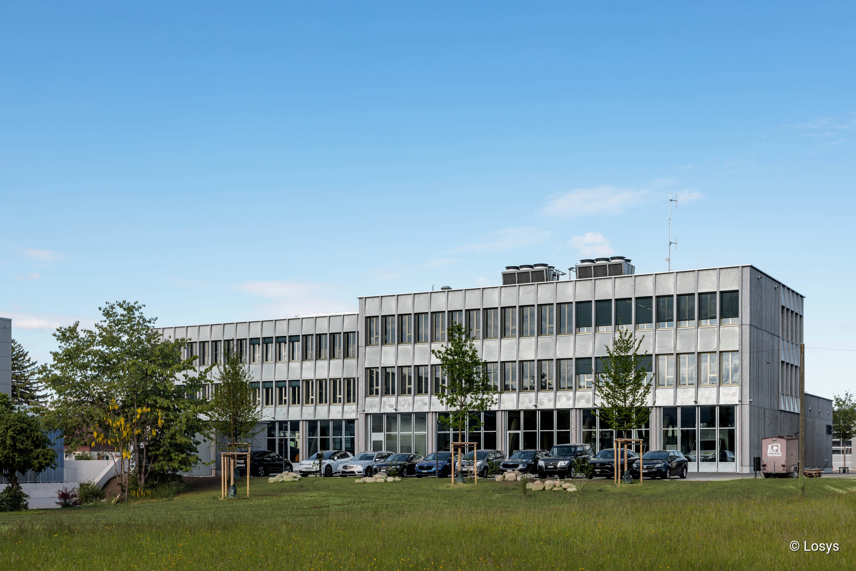Dreigeschossiges Neubau-Werkhof- und Verwaltungsgebäude mit regelmäßiger Beton- und Fensterfassade; im Vordergrund Rasenfläche, neu gepflanzte Bäume und mehrere parkende Autos unter blauem Himmel.