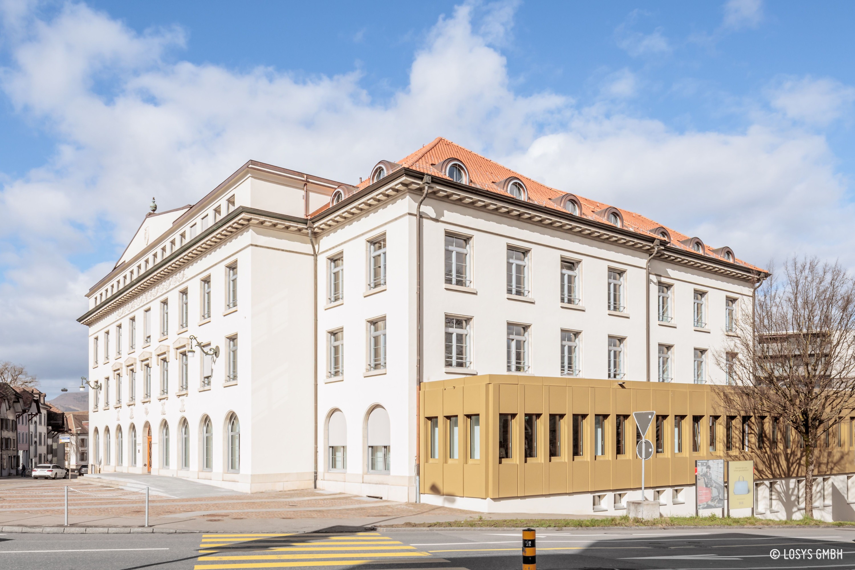 Außenansicht eines sanierten, hellen Eckgebäudes mit rotem Ziegeldach und regelmäßigen Fensterreihen; rechts ein flacher, goldfarbener Anbau mit schmalen Vertikalfenstern. Vordergrund: Straße mit Zebrastreifen und gepflastertem Platz, blauer Himmel, keine Personen.
