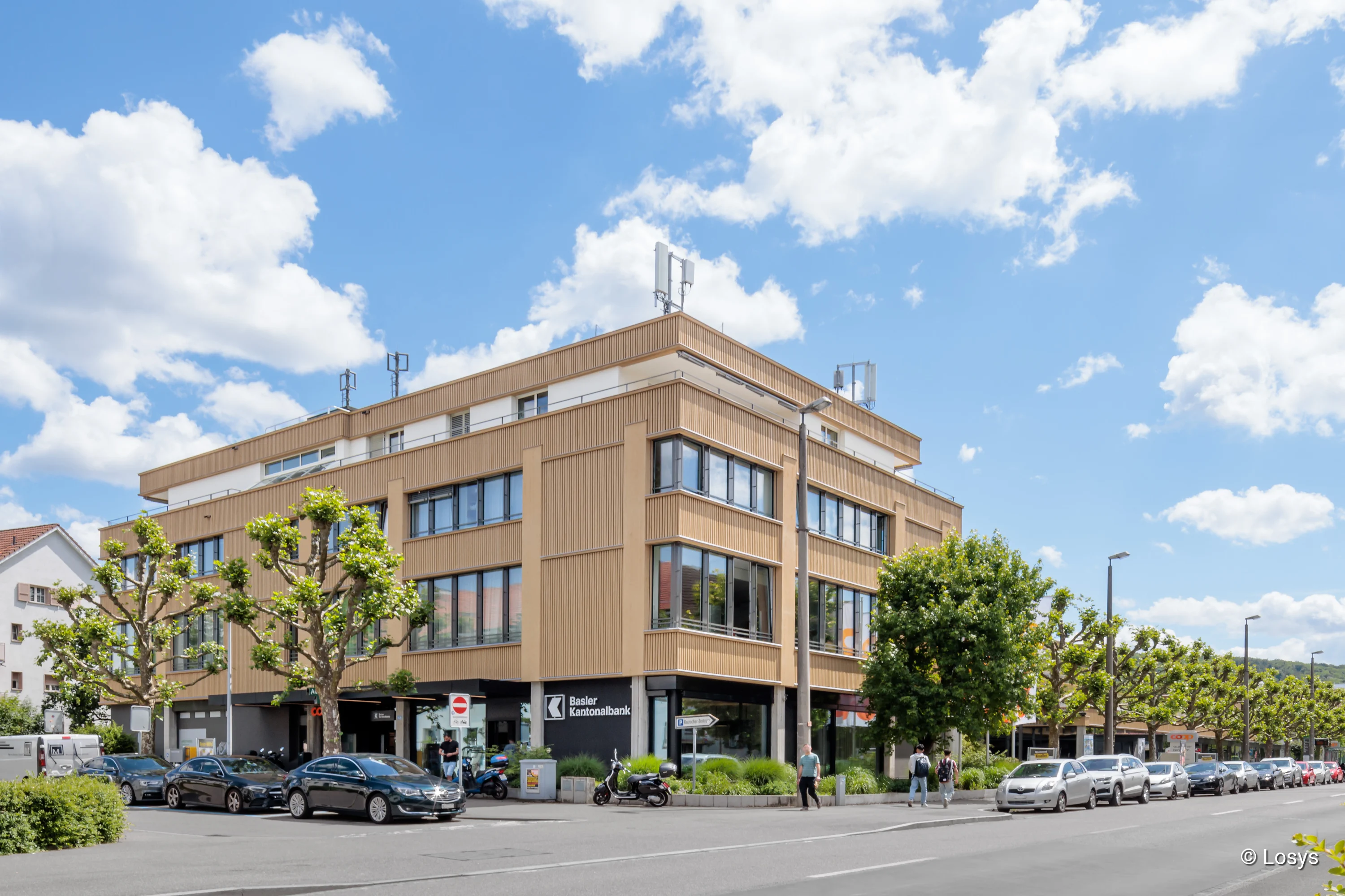 Außenansicht des sanierten Rauracherzentrums in Riehen: viergeschossiges, hellbeiges Gebäude mit vertikaler Holzverkleidung und breiten Fensterbändern. Im Erdgeschoss eine Bankfiliale, vor dem Haus parkende Autos, geschnittene Straßenbäume und mehrere Fußgänger; blauer Himmel mit weißen Wolken.