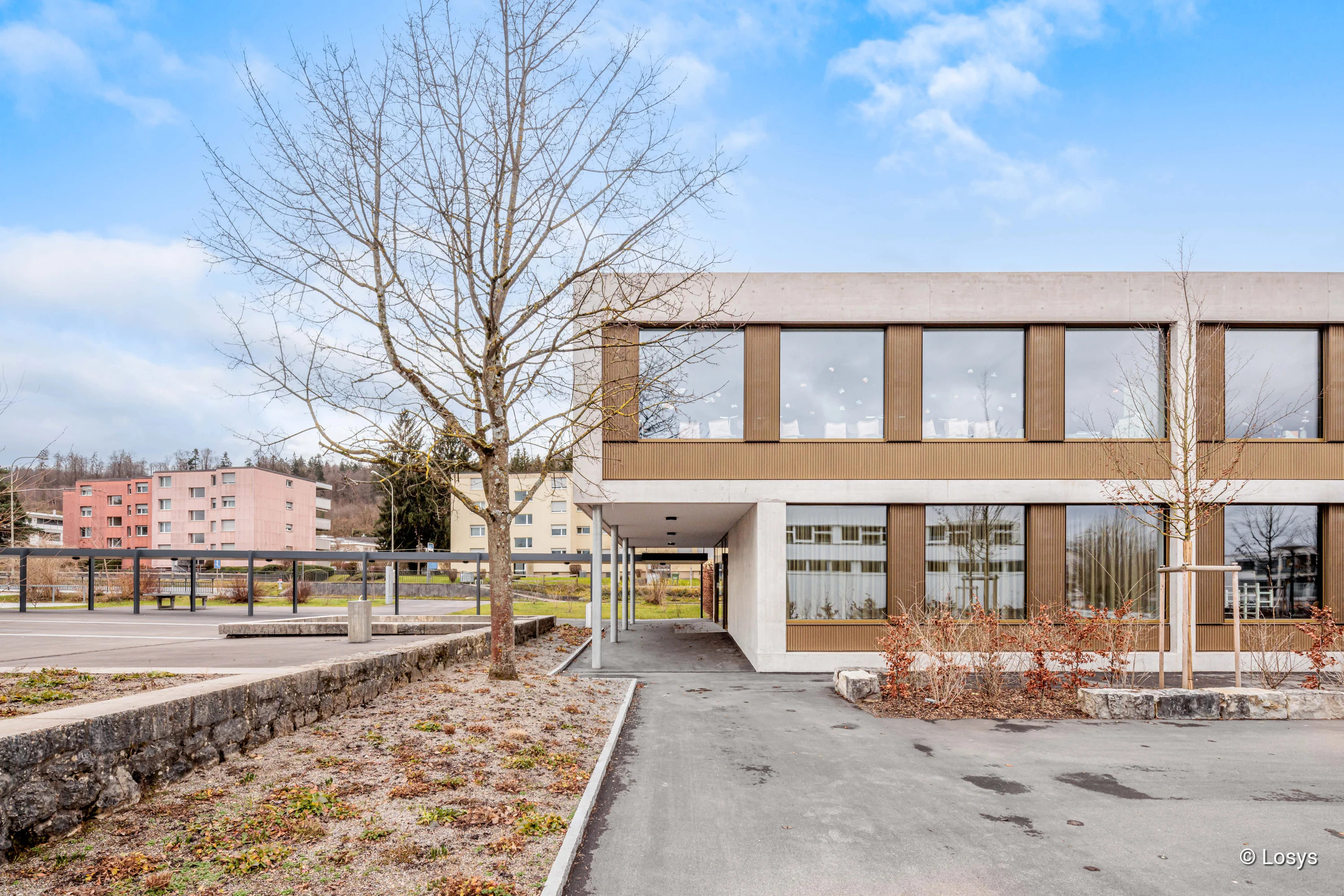 Zweigeschossiges Schulgebäude der Schulanlage Chruezaecher in Regensdorf; Beton- und vertikalholzverkleidete Fassade mit großen rechteckigen Fenstern, überdachter Durchgang mit Stützen, kahler Vorplatz mit Laubbäumen und niedrigen Pflanzbeeten.