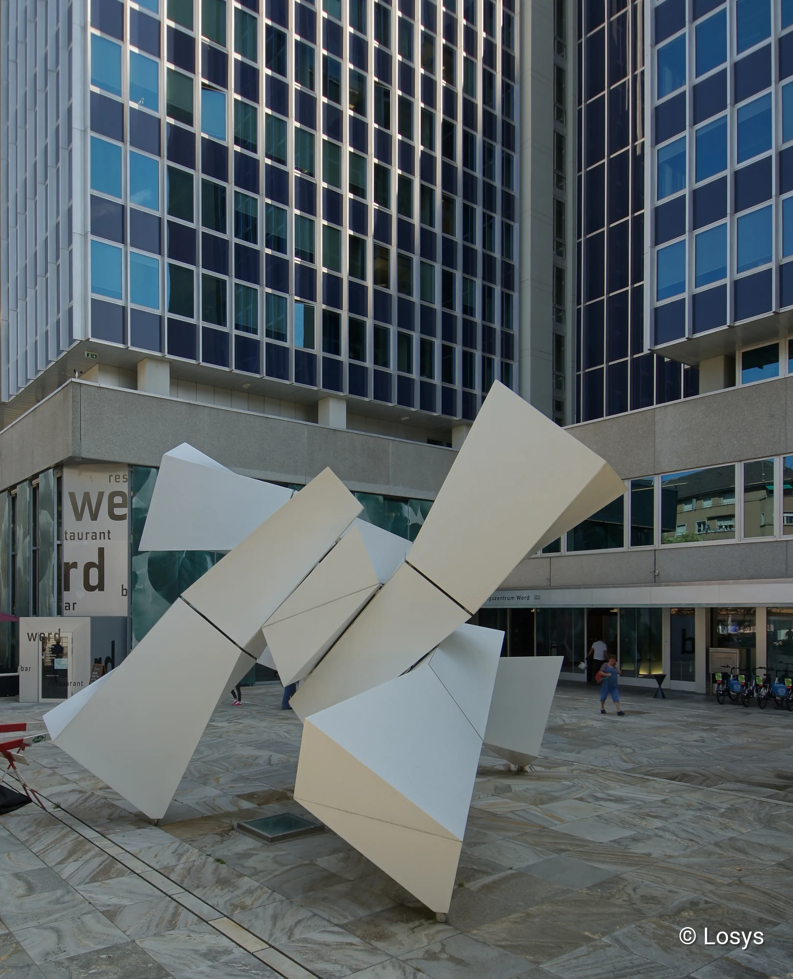 Vorplatz des Verwaltungszentrums Werd (Zürich): im Vordergrund eine große weiße, abstrakte Skulptur aus geometrischen Metalltafeln auf gepflastertem Platz. Im Hintergrund mehrstöckiges Verwaltungsgebäude mit regelmäßiger Rasterfassade aus blau getönten Fenstern, Eingangsbereich, wenigen Passanten und abgestellten Fahrrädern.