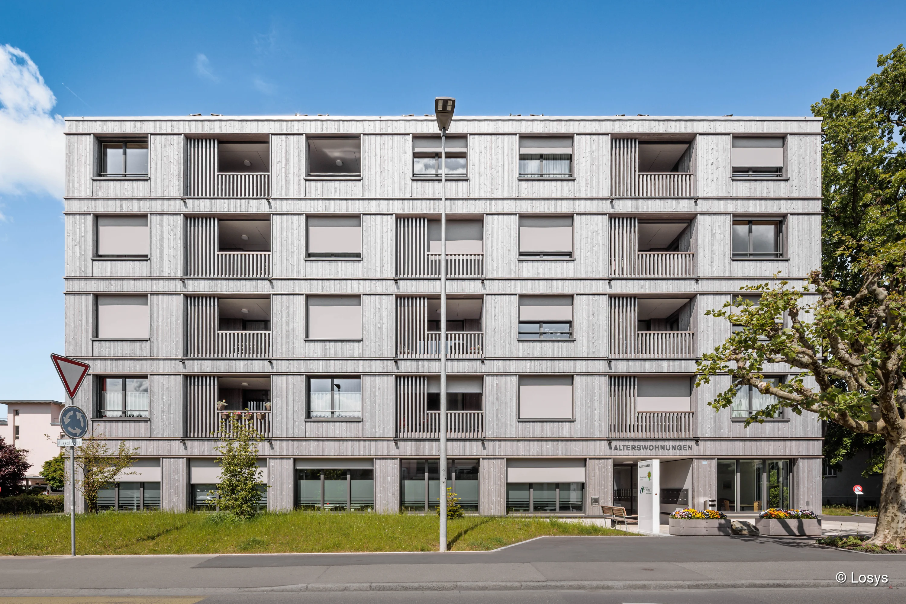 Fassade einer fünfgeschossigen Wohnüberbauung mit hellgrauer, vertikal gegliederter Holzverkleidung. Gleichmäßige Fenster mit heruntergezogenen Sonnenschutz‑Rollos, zurückversetzte Balkone; rechts Haupteingang mit Beschriftung „ALTERS WOHNUNGEN“. Vor dem Haus Rasenfläche, eine Straßenlaterne mittig und ein Verkehrszeichen links, rechts ein beschnittener Baum.