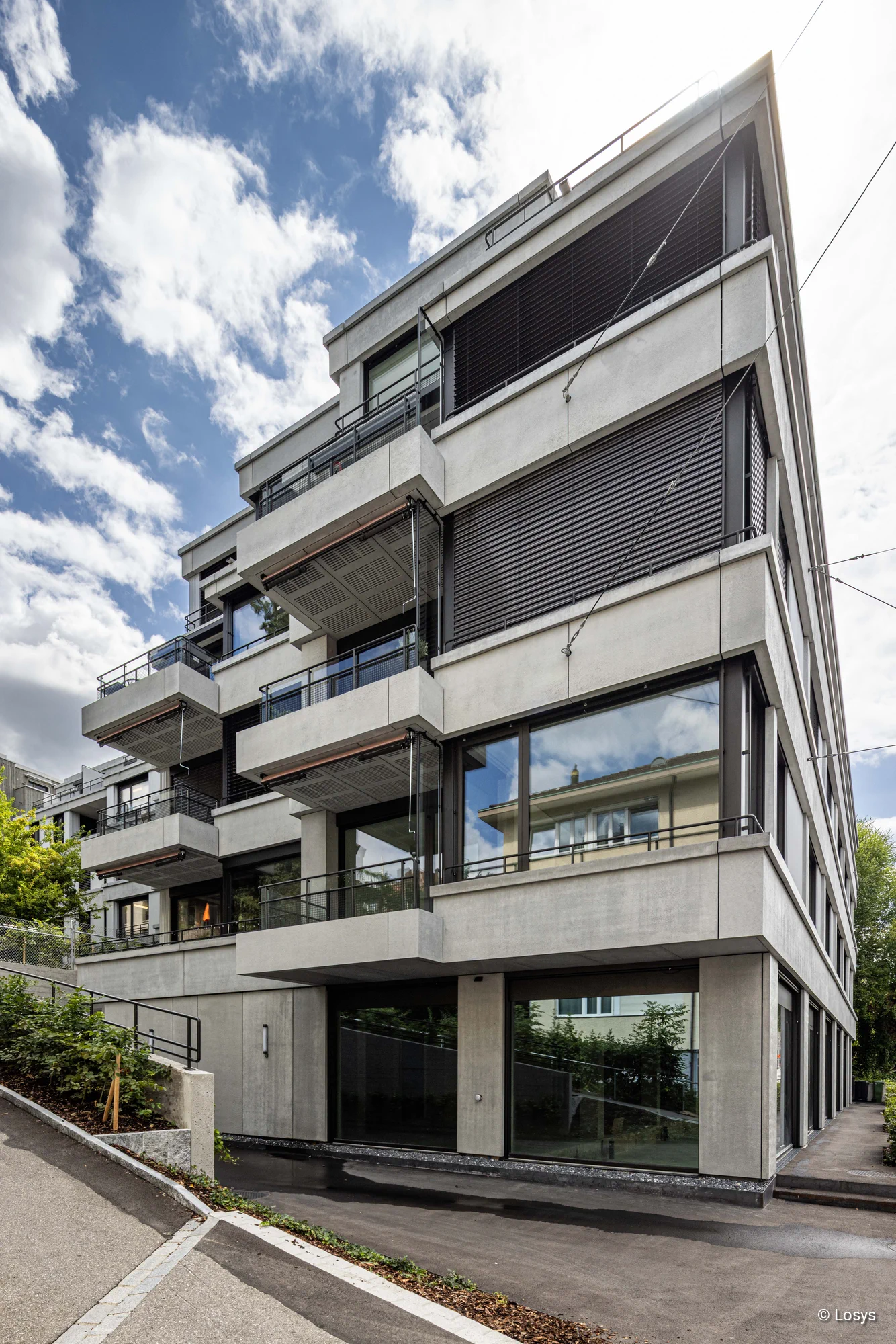 Ecke eines modernen mehrstöckigen Wohnhauses in Zürich: Sichtbetonfassade mit vorstehenden Balkonen, großen Glasfenstern und dunklen Außenjalousien; Metallgeländer, Glasbrüstungen und eine Zufahrt an der Gebäudeecke. Heller Himmel mit Wolken, klarer Kontrast von Beton und dunklen Fensterläden.