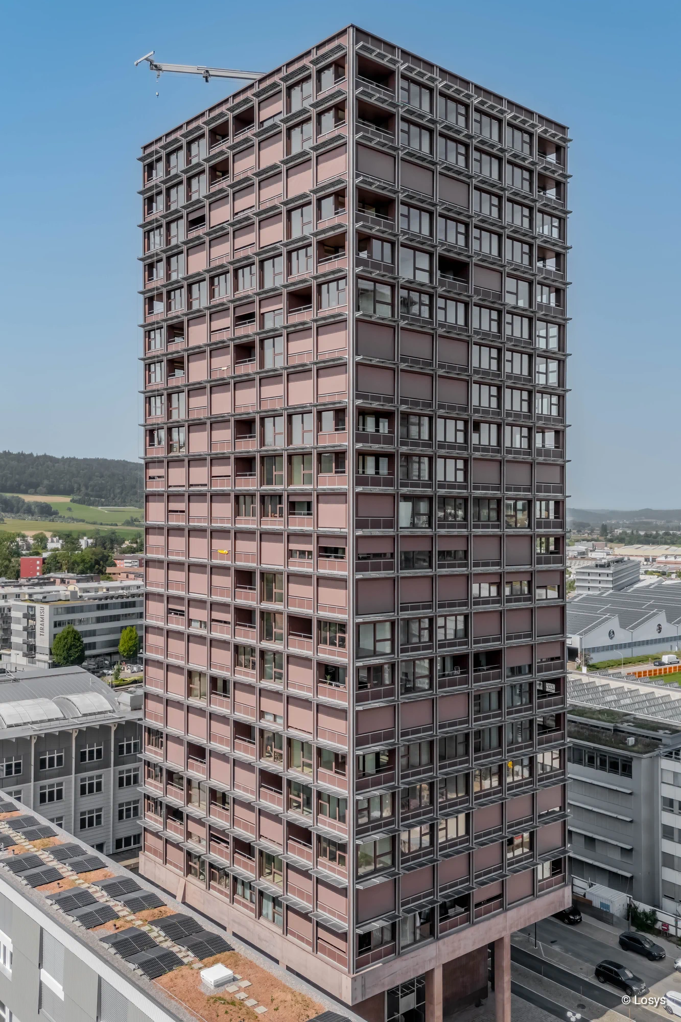 Vogelperspektive auf das Zwhatt-Holzhochhaus in Regensdorf: schlanker Turm mit regelmäßiger Rasterfassade aus Fenstern, Loggien und rosa‑braunen Platten; Kran am Dach und Industrieumfeld mit Solardächern im Vordergrund.